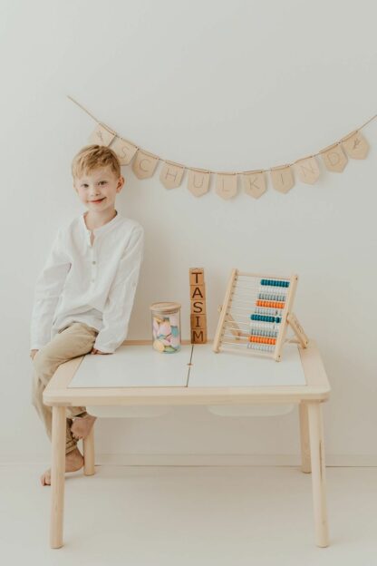 Auf diesem Bild sieht man einen Jungen der auf einem Schreibtisch sitzt mit einer Schulkind Namensgirlande.
