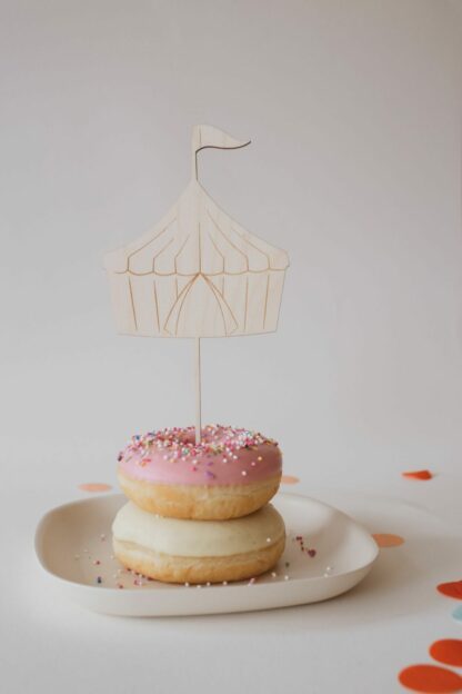 Auf diesem Bild sieht man einen Donut mit einem Cake Topper Zirkuszelt aus Holz.
