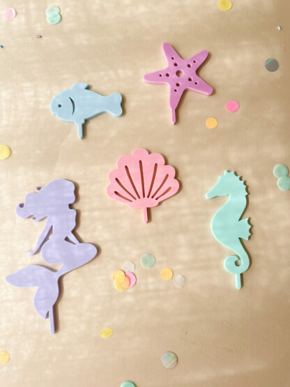 Auf diesem Bild sieht man bunte Stecker in Form von einer Meerjungfrau, Seepferdchen, Muschel, Fisch und Seestern für den Geburtstagskranz aus Holz.