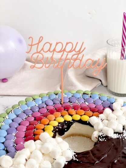Auf diesem Bild sieht man einen Regenbogen Kuchen mit einem bunten Happy Birthday Stecker.