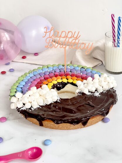 Auf diesem Bild sieht man einen Regenbogen Kuchen mit einem bunten Happy Birthday Stecker und Smarties.